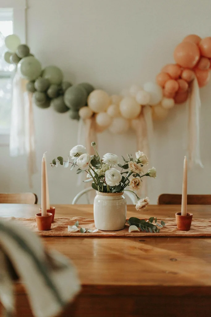 Sage and cream mini balloon garland arch centerpiece over a flower jar on a wood table runner