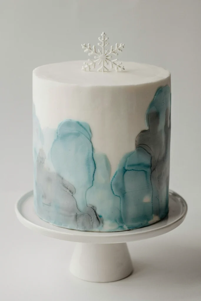 White fondant cake with a soft blended watercolor wash in pale blue and gray, topped with a sugar snowflake