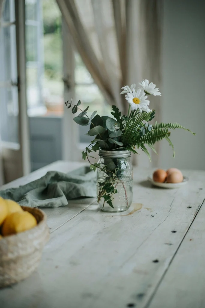 A single large mason jar filled with mixed greenery and white daisies centered on a farmhouse kitchen table