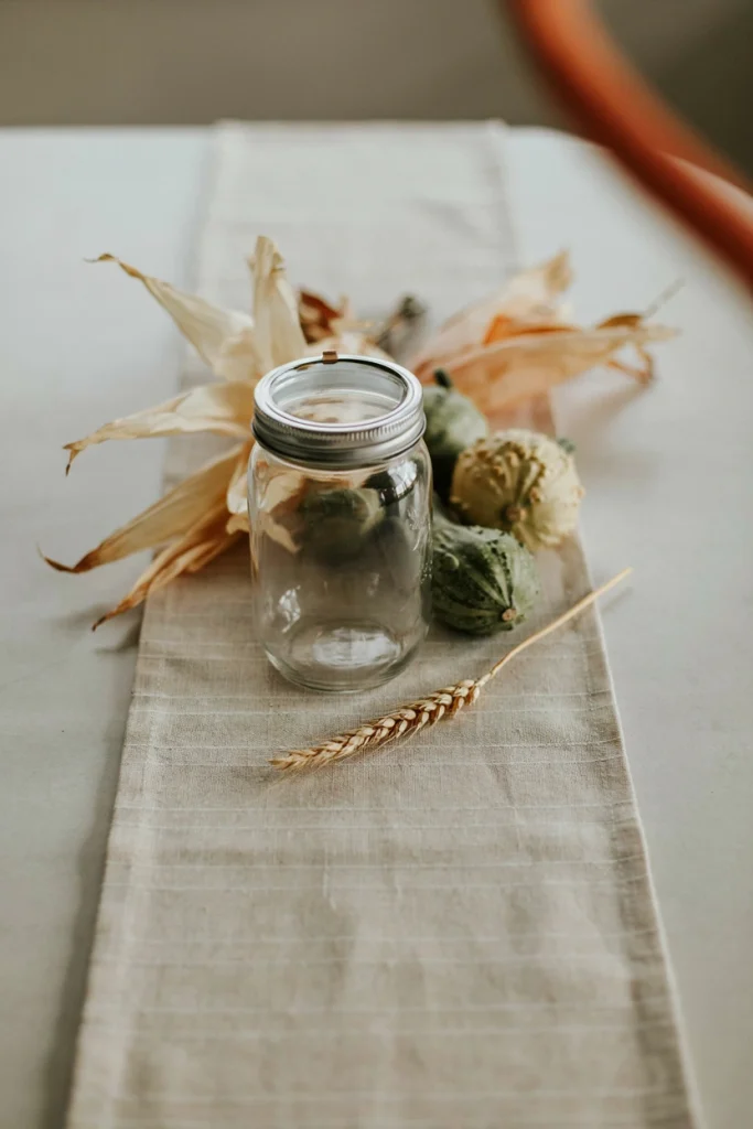 Mason jar surrounded by dried corn husks and small warty gourds in cream and green tones on a woven table runner