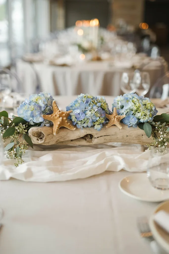 A low centerpiece built on a piece of pale driftwood, with clusters of powder-blue hydrangea and a few dried starfish tucked between the branches