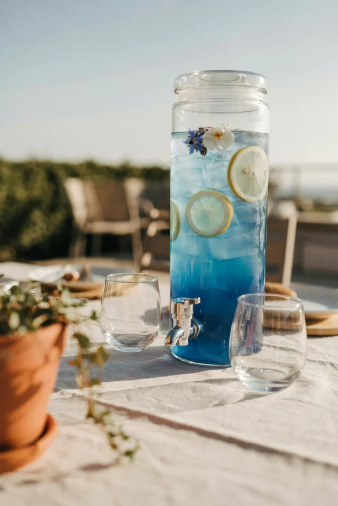 A large glass beverage dispenser filled with a gradient blue mocktail, garnished with lemon wheels and a few edible flowers floating on top